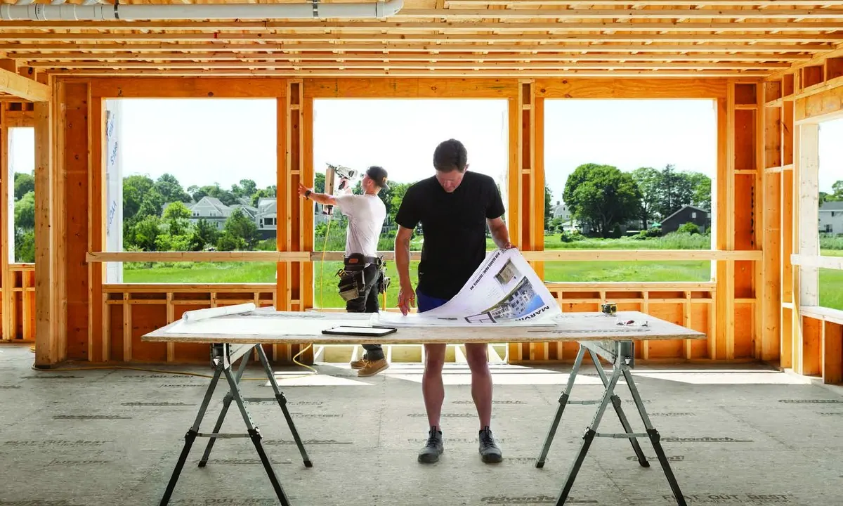 A builder reviewing plans in a framed-out new home with large window openings